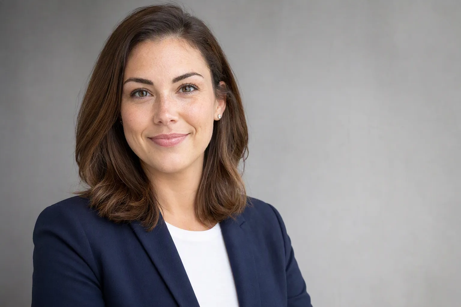 Woman in a navy blazer and white top photographed against a plain grey wall in a professional LinkedIn-style headshot