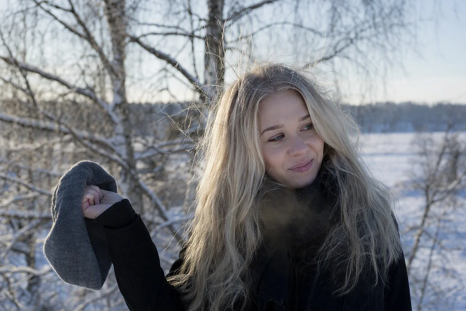 Nainen pitää pipoa kädessään pakkasessa, vaaleat hiukset sähköisinä ja pörröisinä lumisessa maisemassa