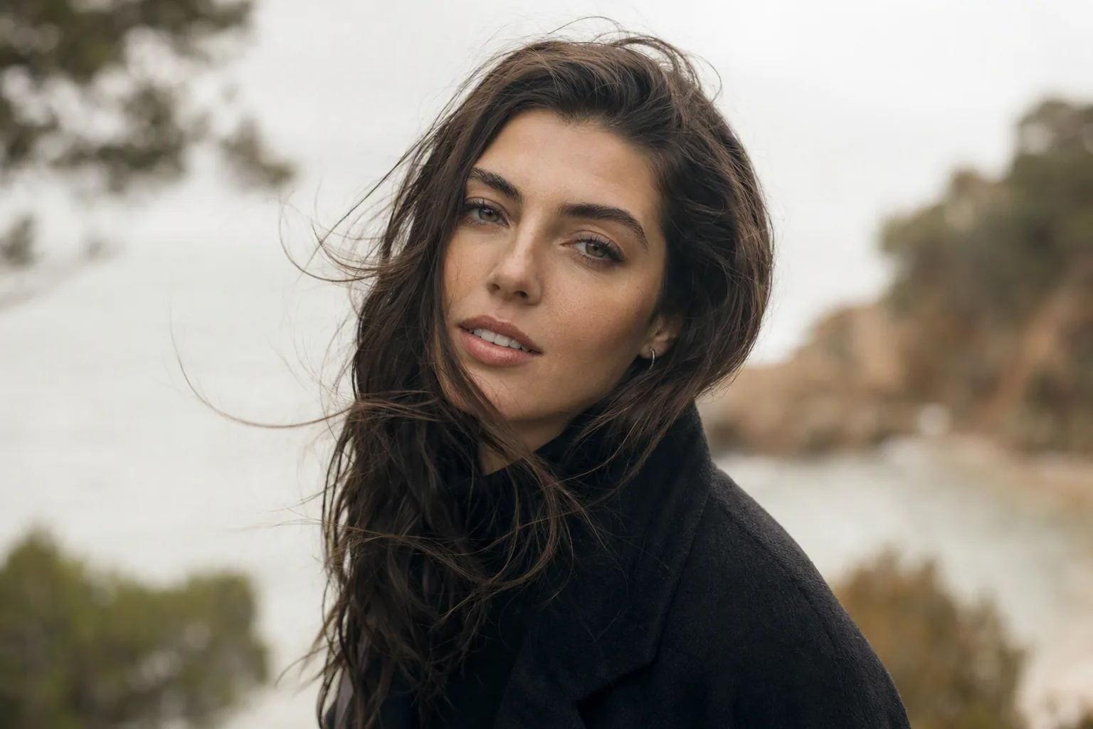 Femme aux longs cheveux bruns décoiffés par le vent, manteau sombre, bord de mer