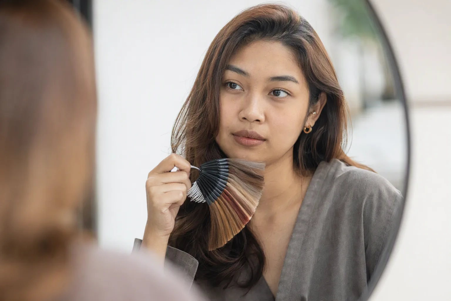 Wanita di depan cermin bulat memegang kipas sampel warna rambut di sisi rambutnya