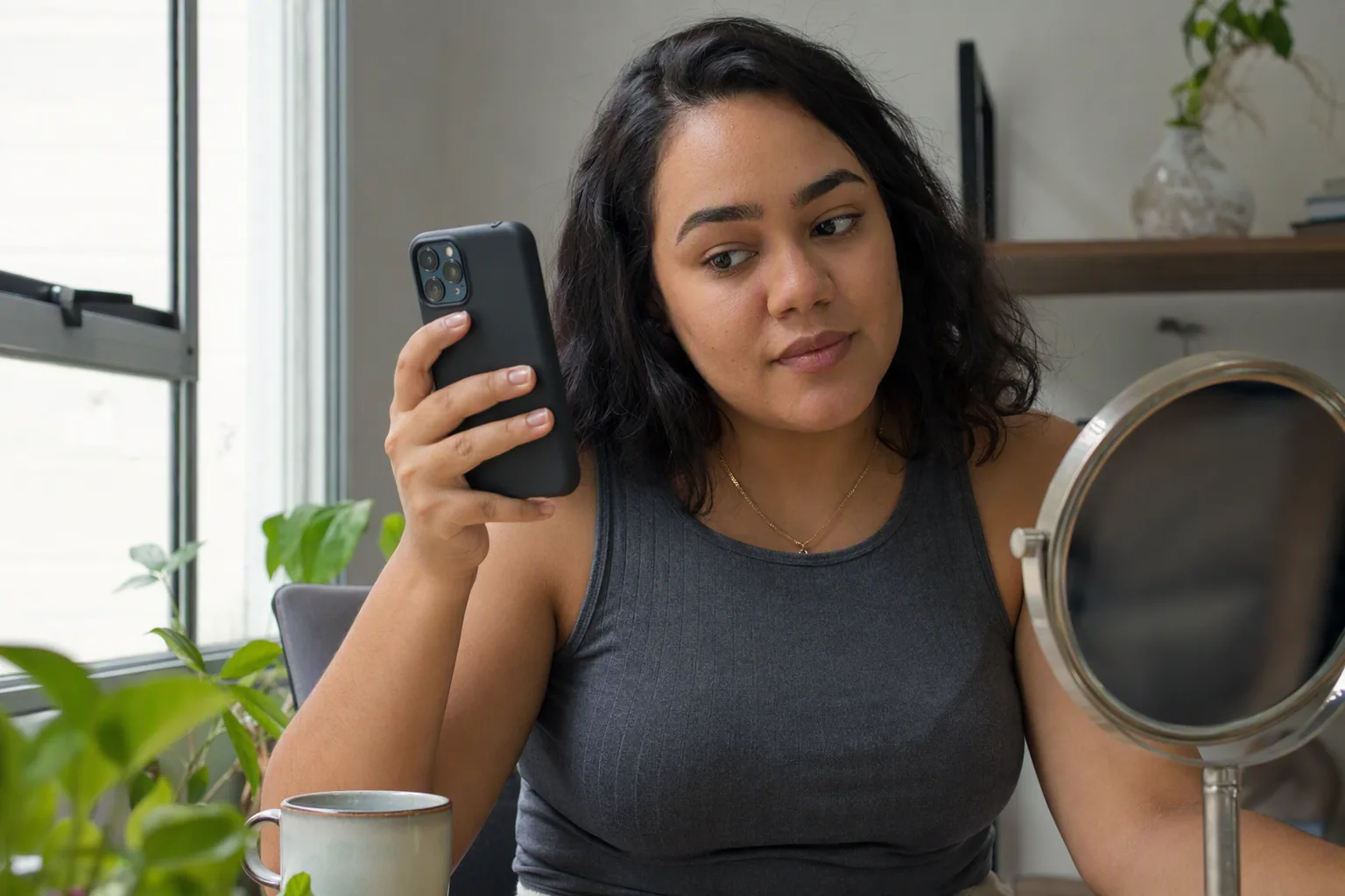 Mulher sentada perto da janela segurando o celular e olhando para um espelho de mesa